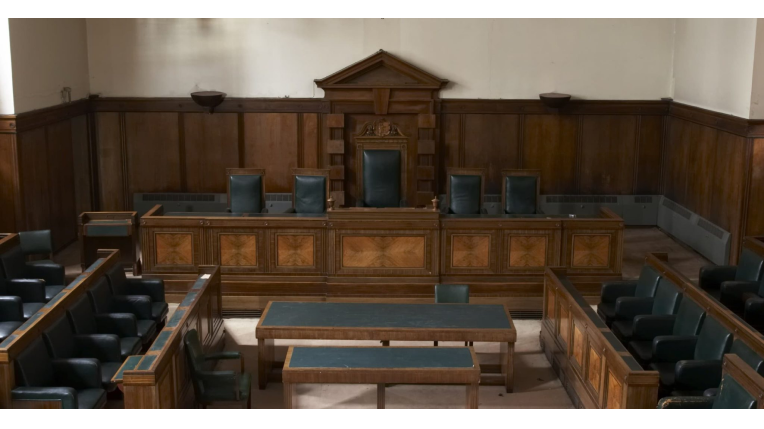 The image shows a courtroom interior. It depicts a formal setting with a raised platform and a large wooden bench at the front. There are rows of chairs on either side, suggesting seating for lawyers, defendants, and possibly the public. Navigating drug charges? Our experienced drug defense lawyers can help.